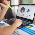 Person analyzing financial charts and graphs on a laptop with colorful documents, showcasing market analysis.